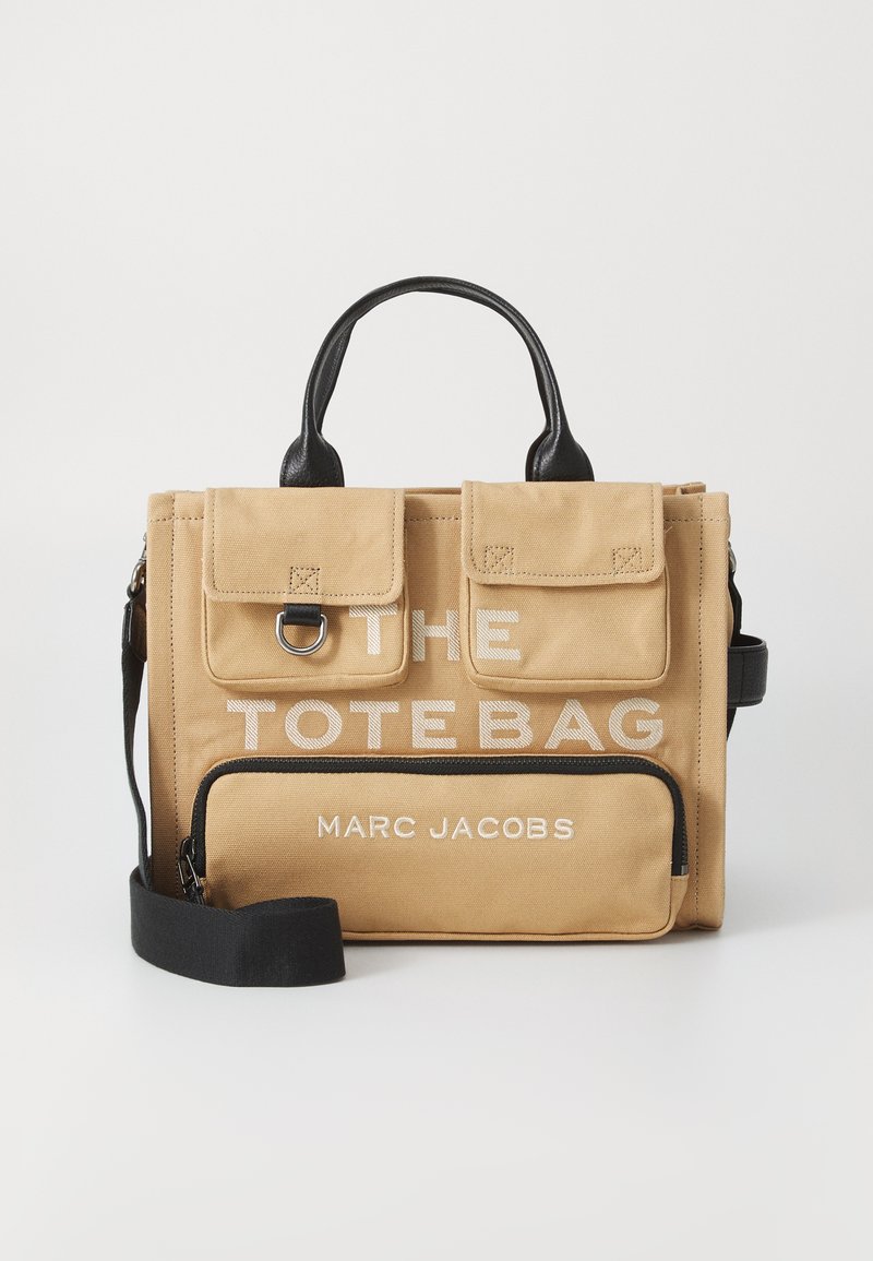 Beeži ristkülikukujuline kott mustade käepidemete ja rihmaga, kaks eesmist klapptaskut, esiline tõmblukukott, tekst "THE TOTE BAG" ja "MARC JACOBS".
