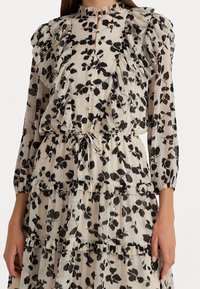 Blommönstrad klänning med volangkrage, långa ärmar och flerlagersdesign. Tyget är lätt och har en genomskinlig, texturerad yta i kräm och svart.