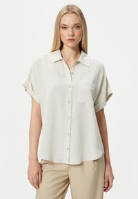 Camisa de manga corta color beige claro en un tejido texturizado, con un frente abotonado y un solo bolsillo en el pecho, y un corte relajado.