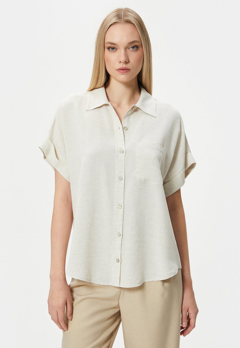 Camisa de manga corta color beige claro en un tejido texturizado, con un frente abotonado y un solo bolsillo en el pecho, y un corte relajado.