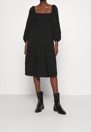 Schwarzes Midikleid mit Puffärmeln, quadratischem Ausschnitt und Raffung; verfügt über einen gestuften Rock. Kombination mit schwarzen Knöchelstiefeln.