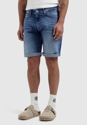 Man die opgerolde blauwe denimshorts draagt, witte sokken met zwarte letters, beige suède instapschoenen en gouden ringen aan de vingers.