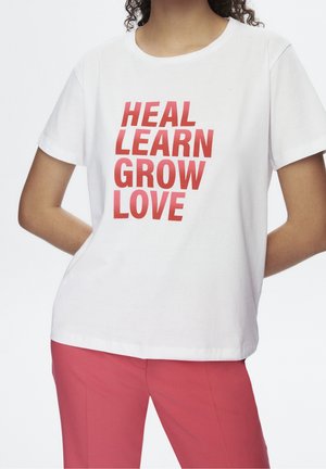 Person wearing a white T-shirt with red text "HEAL LEARN GROW LOVE" paired with pink pants, hands resting behind back.