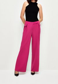 Fuchsiové široké kalhoty s vysokým pasem a skladbami vpředu, doplněné černým topem bez rukávů a černými špičatými podpatky.