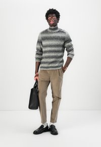 Pull-over à col roulé gris rayé, pantalon beige plissé, mocassins noirs, tenant un sac fourre-tout noir ; design minimaliste, texture lisse.