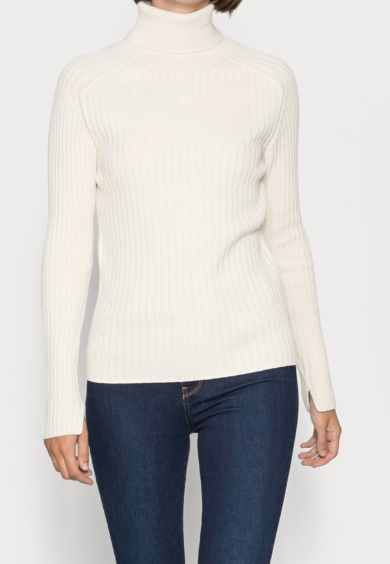 Femme portant un pull à col roulé côtelé crème avec des poignets avec trous pour le pouce et un jean bleu foncé sur un fond uni.