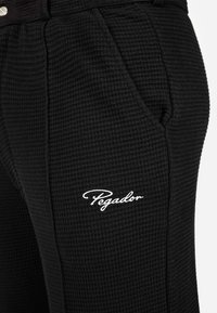 Fekete, texturált nadrág kockás mintával, oldalzseb közelében fehér hímzett "Pegador" logóval.