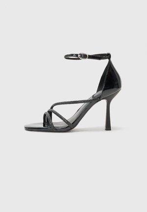 Črne lakirane sandale na visoki peti z naramnicami, ki imajo križaste trakove in elegantno konico.