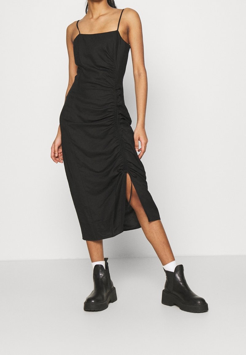 Robe midi noire avec bretelles spaghetti, détails froncés sur le côté et une fente haute. Portée avec des bottines noires épaisses et des chaussettes blanches.