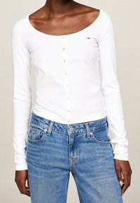 Top blanco de manga larga hecho de tela de canalé, con escote en forma de barco y cierres de botones en la parte delantera, combinado con jeans de mezclilla azul claro.