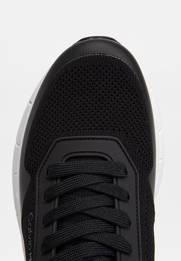 Baskets noires avec un dessus en tricot texturé, un capuchon de orteil lisse noir, des lacets noirs et une semelle blanche contrastante. Présente un accent de logo sur le côté.