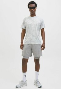 Harmaa lyhythihainen urheiluvaate, jossa on hillitty kuvio, yhdistettynä samanvärisiin harmasin shortsien kanssa. Mallilla on tummat aurinkolasit ja hopeiset juoksukengät.