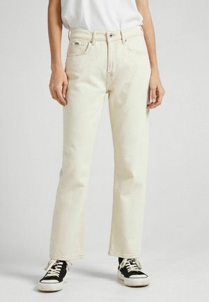 Person iført off-white jeans med lige ben og sort-hvide sneakers, stående mod en ensfarvet lys baggrund.