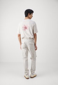 Camiseta de color beige claro con un gráfico floral y texto en la parte posterior, combinada con pantalones cargo beige claro y zapatillas blancas con acentos amarillos.