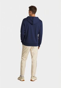 Felpa con cappuccio blu navy con finitura testurizzata, indossata con pantaloni chiari e sneakers color sabbia. Dettagli delle cuciture visibili sulle maniche.