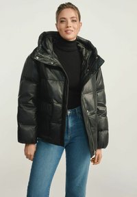 Czarna kurtka puchowa o błyszczącej powierzchni, z zapi�ęciem na zamek, dużym kapturem i przednimi kieszeniami. Nosi się ją na czarnym golfie i niebieskich dżinsach.