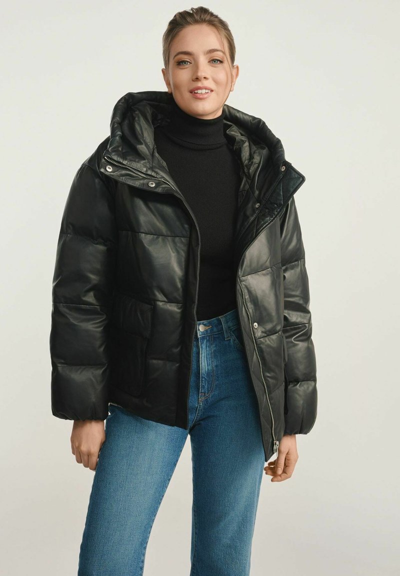 Czarna kurtka puchowa o błyszczącej powierzchni, z zapięciem na zamek, dużym kapturem i przednimi kieszeniami. Nosi się ją na czarnym golfie i niebieskich dżinsach.