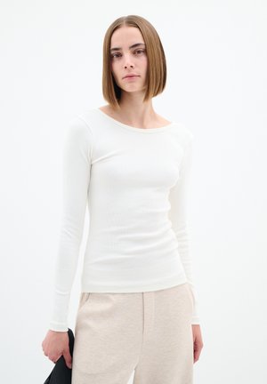 Langärmliges, geripptes Oberteil in cremiger Farbe mit weitem Ausschnitt. Glatte Textur, figurbetontes Design, getragen mit hellbeigen Hosen und einer schwarzen Clutch.