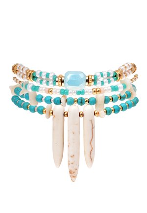 Bracelet multi-couches avec des perles blanches et turquoise, des accents dorés, et trois pointes blanches allongées. Des matériaux texturés créent un contraste.