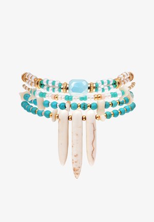 Bracelet multi-couches avec des perles blanches et turquoise, des accents dorés, et trois pointes blanches allongées. Des matériaux texturés créent un contraste.