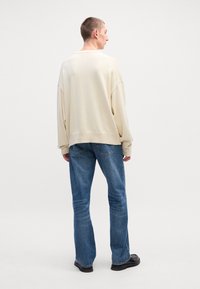 Κρέμα oversized πουλόβερ με χαμηλωμένα ώμους, συνδυασμένο με ανοιχτό μπλε ίσια τζιν και μαύρα παπούτσια, προβολή από πίσω.