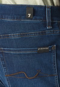 Jeans in denim blu scuro con un'etichetta nera con il numero "7" e un design della tasca cucito con filo marrone. L'etichetta riporta la scritta "per tutta l'umanità."