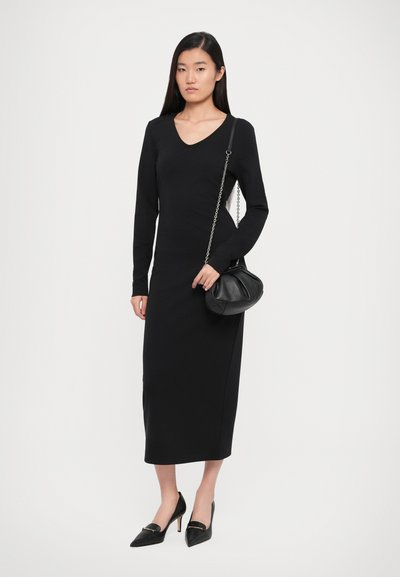 Black long-sleeve dress with a V-neckline, fitted shape, and knee-length hem, paired with a black quilted handbag and pointed shoes.
