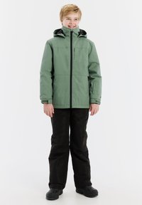 Grüne wasserfeste Jacke mit schwarzen Reißverschlüssen; Kapuzendesign. Tragend mit schwarzen Hosen und schwarzen Stiefeln, stehend vor einem weißen Hintergrund.