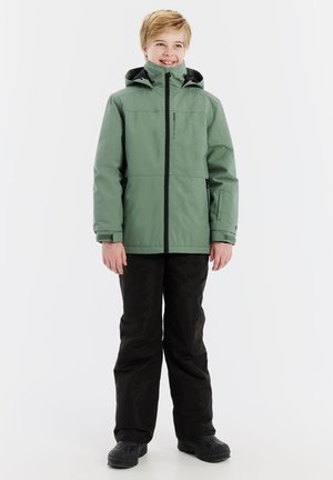 Grüne wasserfeste Jacke mit schwarzen Reißverschlüssen; Kapuzendesign. Tragend mit schwarzen Hosen und schwarzen Stiefeln, stehend vor einem weißen Hintergrund.