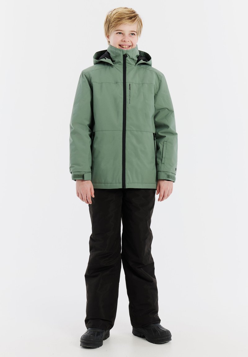 Grüne wasserfeste Jacke mit schwarzen Reißverschlüssen; Kapuzendesign. Tragend mit schwarzen Hosen und schwarzen Stiefeln, stehend vor einem weißen Hintergrund.