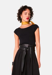 Zwarte aansluitende top met kapmouwen, gecombineerd met een zwarte geplooide rok en een brede leren riem. Een gele stippen sjaal accentueert de halslijn.