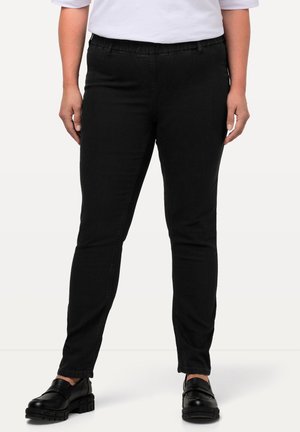 Persona vistiendo pantalones negros ajustados, mocasines negros gruesos y una camiseta blanca de manga corta, de pie frente a un fondo liso.