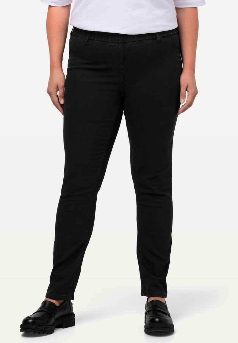 Persoon draagt nauwsluitende zwarte broek, zwarte stevige loafers en een wit shirt met korte mouwen, staand tegen een effen achtergrond.