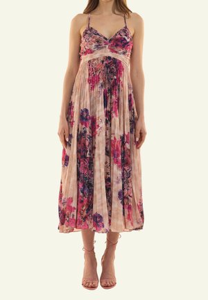 Frau trägt ein ärmelloses, plissiertes Midikleid mit Blumenmuster in Pink-, Lila- und Beigetönen, kombiniert mit nude-farbenen Riemchen-High Heels.