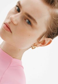 Pendiente de aro dorado con un diseño retorcido, usado en la oreja izquierda. La persona tiene el cabello corto y lleva una blusa rosa ajustada.