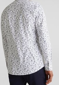 Chemise à manches longues blanche avec un motif floral bleu marine. Elle présente un col classique et des poignets à boutons, fabriquée en tissu léger.