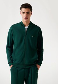 Grøn zip-up træningsjakke lavet af glat stof, med ståkrave, sidelommer og et lille logo i øverste venstre hjørne.