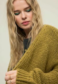 Langer, senfgelber, gestrickter Cardigan mit gerippten Bündchen, getragen über einem dunklen Oberteil. Die Haare sind wellig und fallen seitlich ins Gesicht.