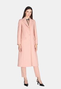 Cappotto di lana rosa con design sartoriale, maniche lunghe, rever a punta, bottoni frontali e due tasche laterali, abbinato a pantaloni beige e décolleté neri.