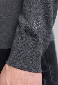 Mörkgrå stickad tröjsleeve med ribbad manschett; har ett subtilt ljusgrått emblem som liknar en blommönster. Slät textur, åtsittande design.