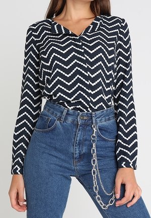 Kvinde iført en langeærmet sort-hvid zigzag-mønster skjorte stoppet ned i højtaljede blå jeans med en sølvkæde som tilbehør.