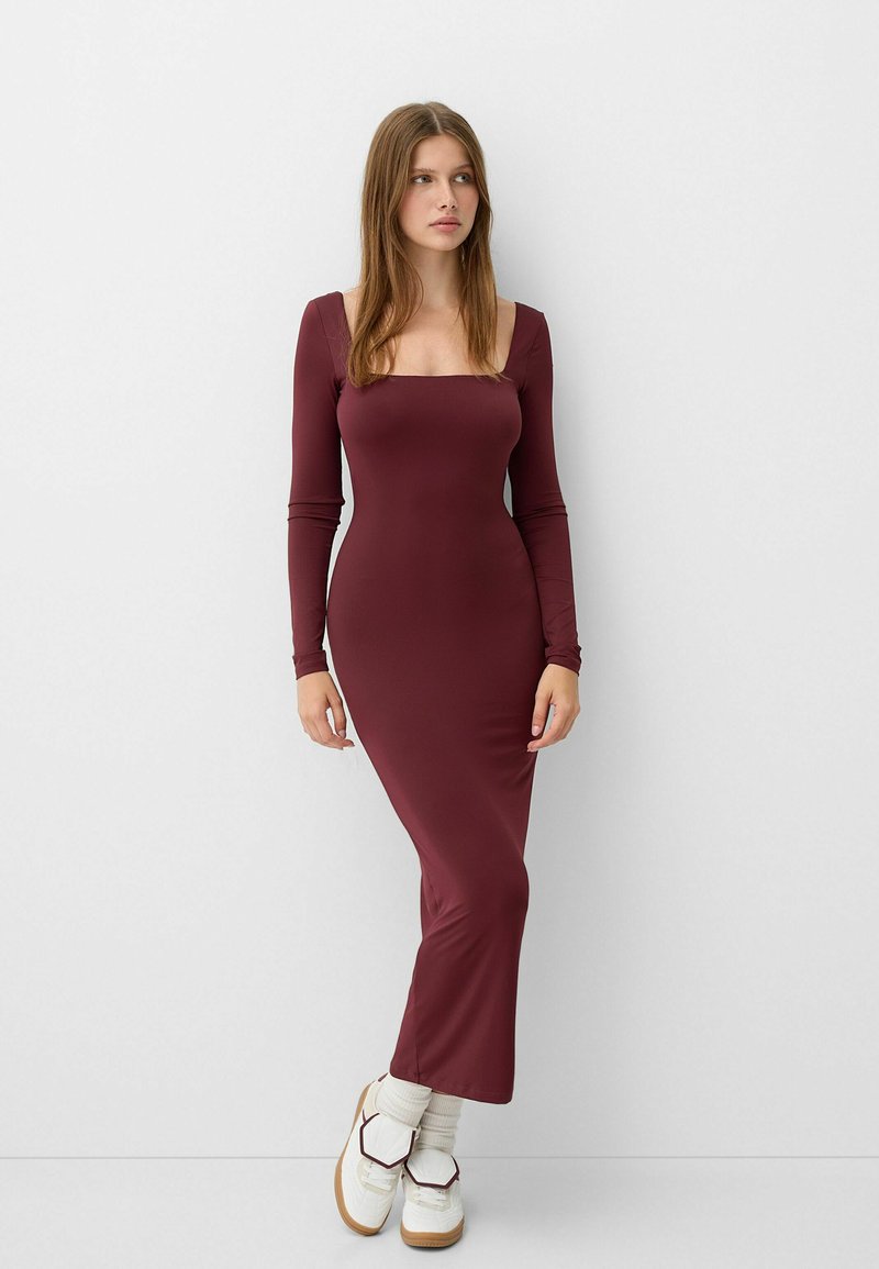Abito lungo bordeaux a maniche lunghe con scollatura quadrata, silhouette aderente, realizzato in tessuto liscio, indossato con sneakers bianche e calzini crema.