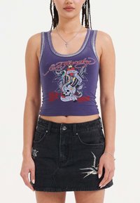 Top corto senza maniche viola con un design grafico che presenta un teschio, serpenti e un uccello. Abbinato a una gonna di denim nera a vita alta con ricami.