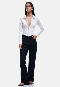 Biała, karbowana bluzka zapinana na zamek z kołnierzykiem zestawiona z ciemnoniebieskimi spodniami typu wide-leg. Modelka nosi czarne sandały; minimalistyczny design z czystymi liniami.