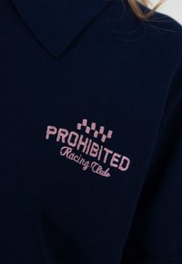 Chemise bleu marine à col avec texte brodé rose "PROHIBITED Racing Club" et un motif de drapeau à damiers rose au-dessus du texte.
