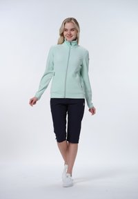 Mintgrön zip-jacka med hög krage, tillsammans med marinblå knälånga shorts och vita sneakers, mot en plain bakgrund.
