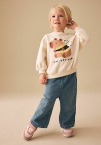 Sweat-shirt crème avec un graphisme d'ours et une abeille, associé à un pantalon en denim bleu ample et des baskets roses avec des accents blancs.