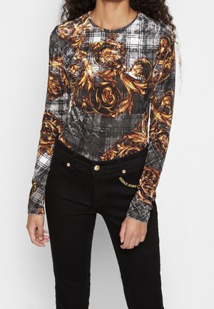 Vrouw draagt een shirt met lange mouwen in zwart en grijs ruitpatroon en goud barokprint, gecombineerd met een zwarte spijkerbroek met gouden knopen en logodetail.