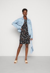 Casaco de ganga sobre um vestido preto floral com padrões em laranja, azul e branco. Sapatos de salto bloco abertos em bege e uma mala azul clara.