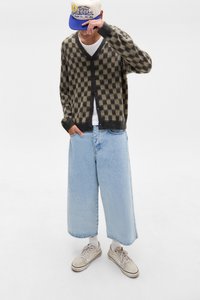 Cardigan în carouri, maro și bej, cu nasturi, fit oversized. Pantaloni din denim cu picioare largi de culoare albastru deschis și teniși bej cu ciorapi albi.